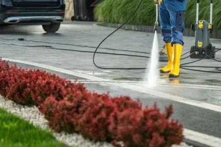 A person in yellow boots pressure washes a paved driveway. Red bushes line the side of the driveway, with a car parked in the background. Location: Fort Lauderdale, United States